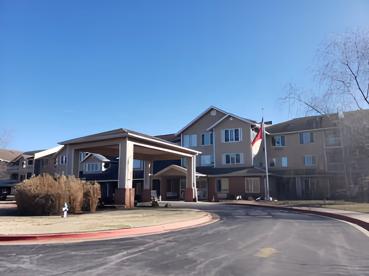 Front entrance of a multi-story senior living building with a covered porte-cochere, flagpole, and clear blue sky.