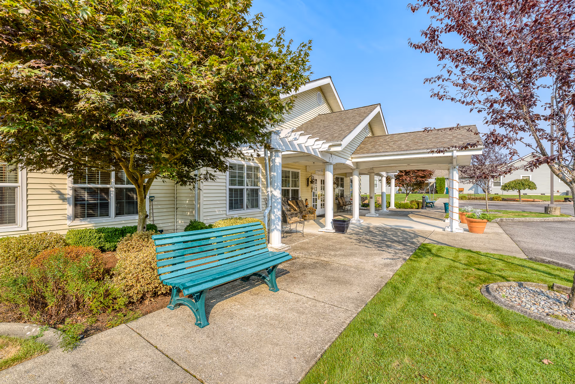 Outdoor view of the entrance area of Cascade Place Assisted Living, Memory Care facility featuring a covered walkway with white columns, a green bench, trees, shrubs, and a well-maintained lawn under a clear blue sky.