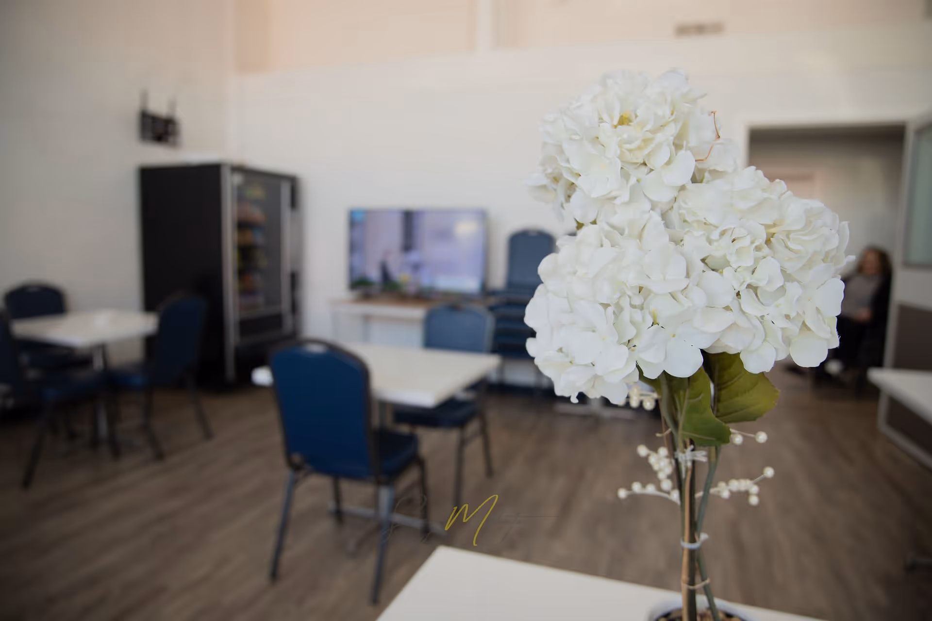A room with several tables and blue chairs, a vending machine against the wall, a television on a stand, and a bouquet of white flowers in the foreground. The room has wood flooring and white walls.