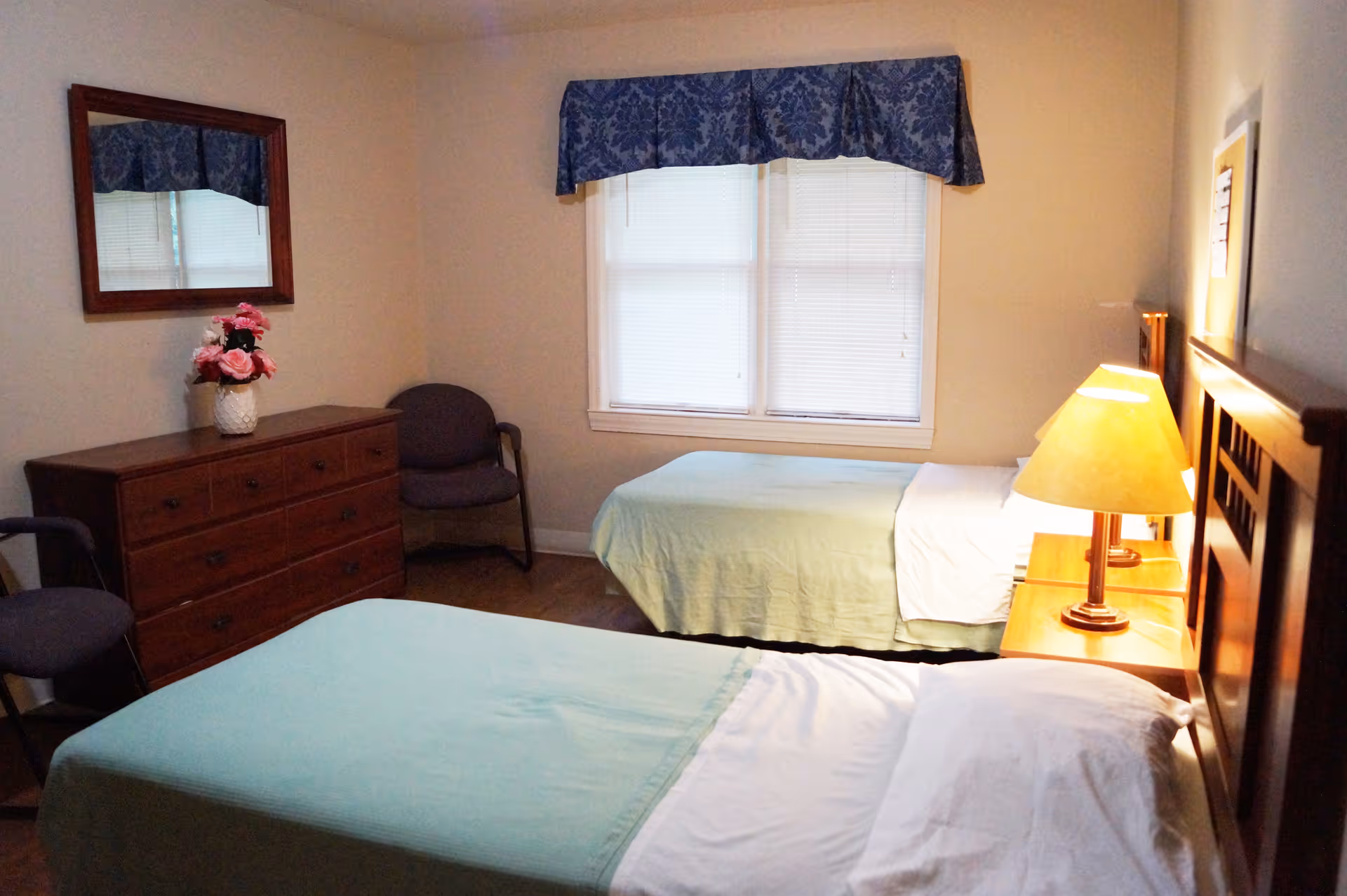 Small senior living bedroom with two twin beds, wooden dresser, chairs, and a window with blinds.