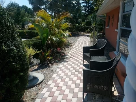 Outdoor patio area with a checkered brick walkway, two black wicker chairs, and various green plants and trees surrounding the path next to a building with orange walls and windows.