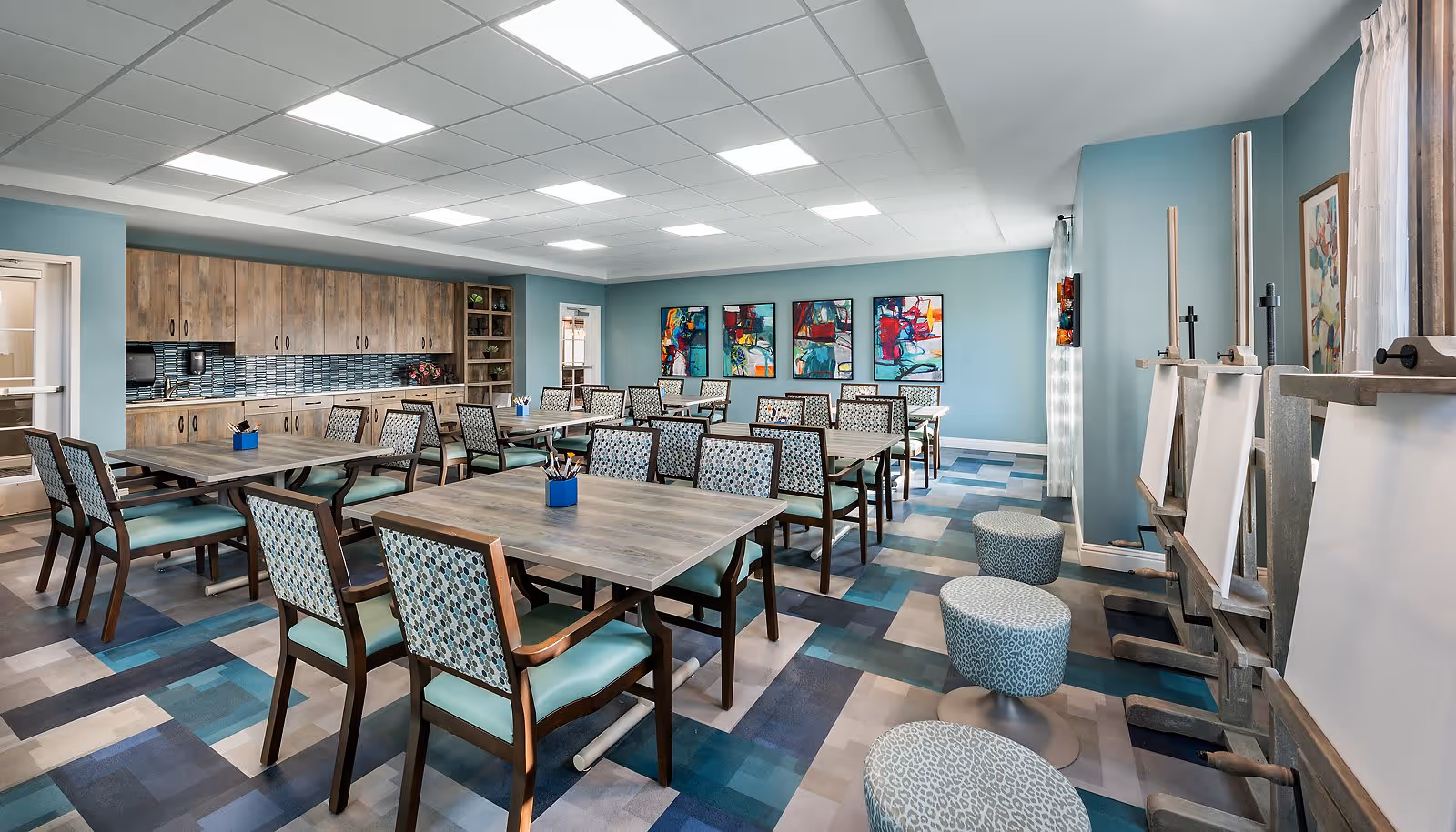 A spacious activity room with multiple tables and chairs arranged neatly. The room has light blue walls adorned with colorful abstract paintings. There is a kitchenette area with wooden cabinets and a tiled backsplash. On the right side, there are easels and round patterned stools near a window with sheer curtains, suggesting an art or craft space.