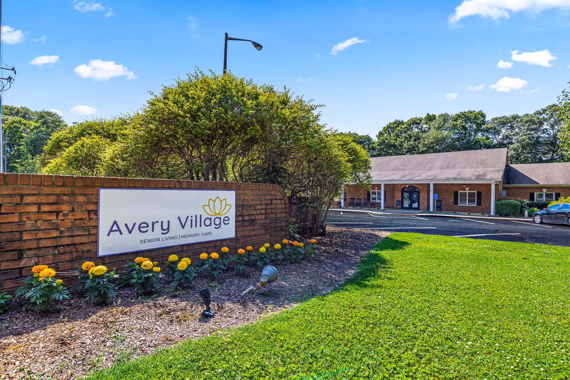 Exterior view of Avery Village Assisted Living and Memory Care facility with a brick sign in the foreground surrounded by yellow and orange flowers, green grass, and bushes. The building is a single-story structure with a sloped roof and a parking area in front under a clear blue sky.