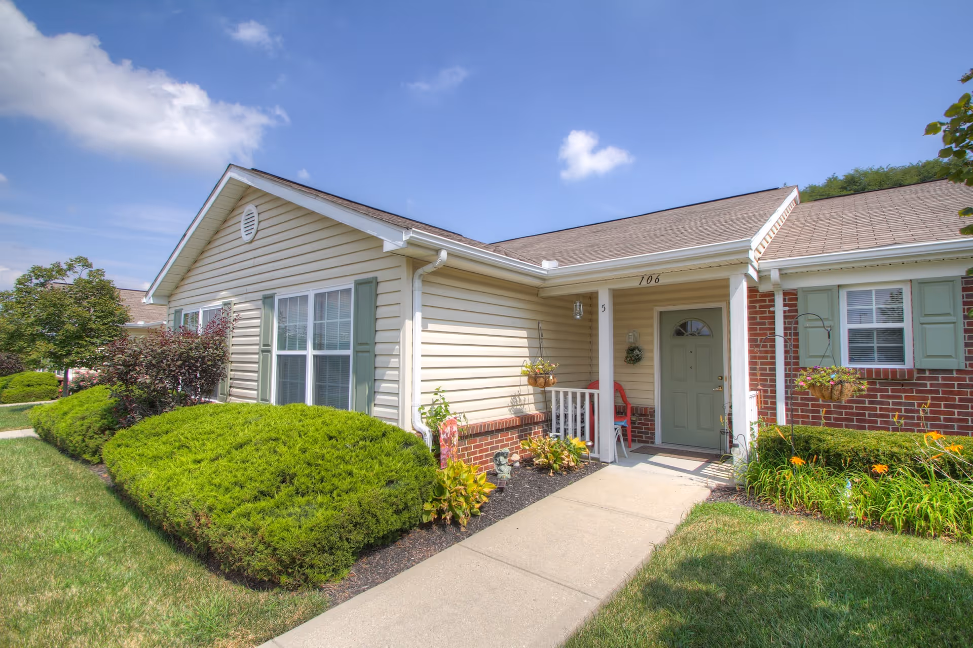 Exterior view of a single-story residential unit at Riverview Bluffs with beige siding, green shutters, a green front door, and a small covered porch. The walkway is bordered by well-maintained green bushes and flowering plants under a partly cloudy blue sky.