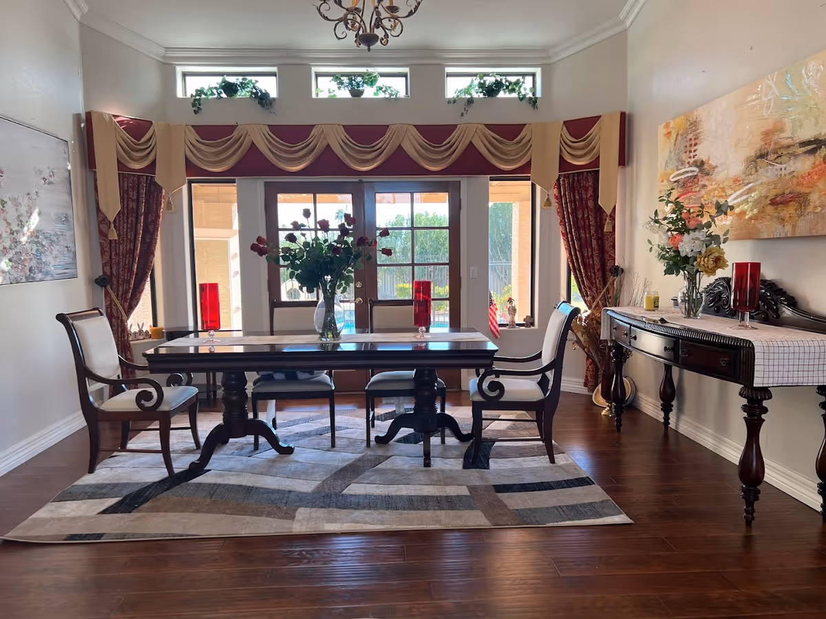 A formal dining room with a dark wooden table and six upholstered chairs. The table has a vase with red roses and two red candle holders. Behind the table are glass doors leading outside, with decorative curtains and valances above. There is a sideboard on the right with a floral arrangement and a large abstract painting hanging above it. The floor is wooden with a patterned area rug under the table.