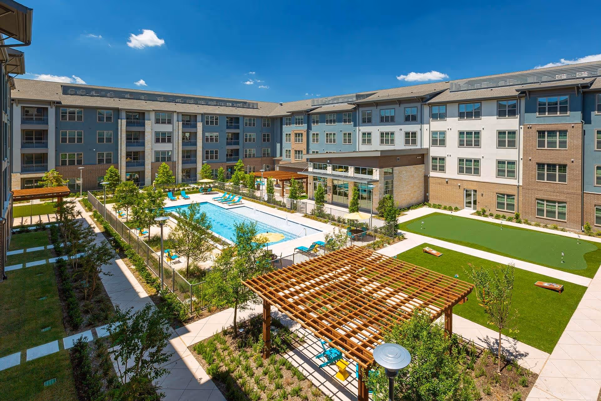 Outdoor courtyard area of The Orchards Market Plaza featuring a swimming pool with lounge chairs, a putting green, landscaped gardens, walkways, and a wooden pergola with seating under a clear blue sky.