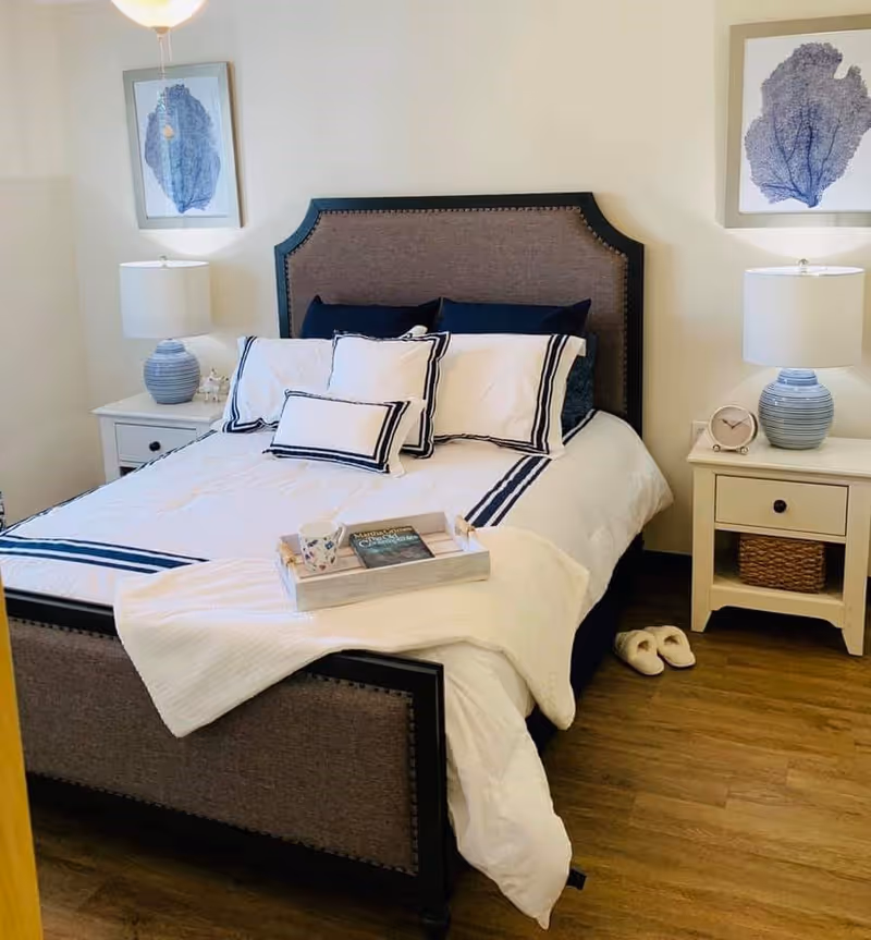 A neatly made bed with white and navy blue bedding in a bedroom. The bed has a padded headboard and footboard with dark trim. On the bed is a tray holding a mug and a book. There are two white nightstands on either side of the bed, each with a blue-striped lamp and decorative items. A pair of white slippers is placed on the wooden floor next to the right nightstand. Two framed blue coral prints hang on the wall above each nightstand.