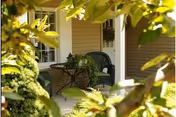 A cozy outdoor patio area with a black metal chair and a small round table holding a potted plant, surrounded by green foliage and partially shaded by tree branches.