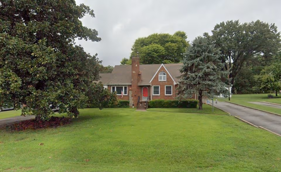 A brick house with a red front door, surrounded by green grass and trees under a cloudy sky.