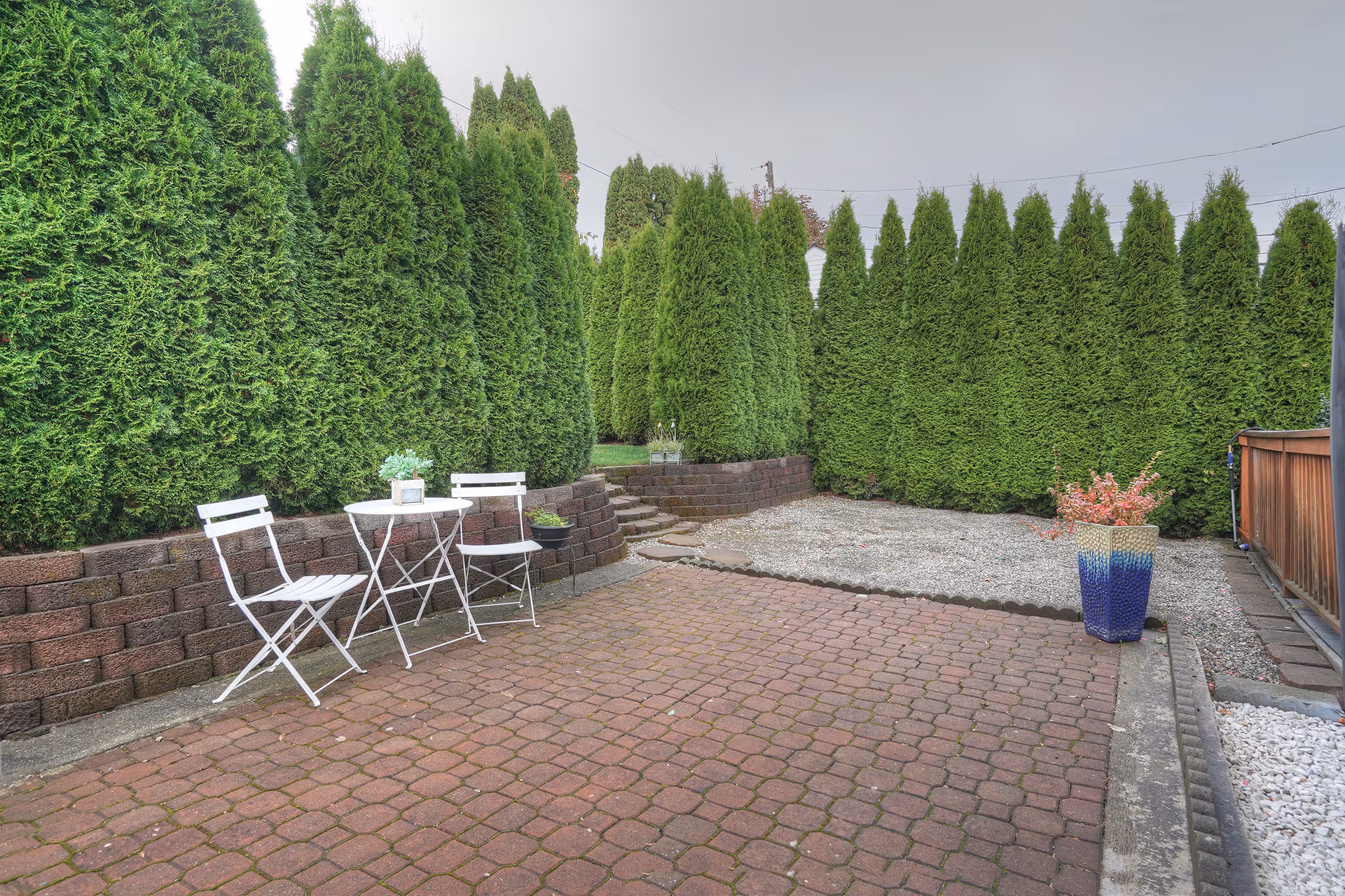 Paved patio with a white bistro table and chairs beside tall evergreen hedges, a gravel area, and a blue planter.