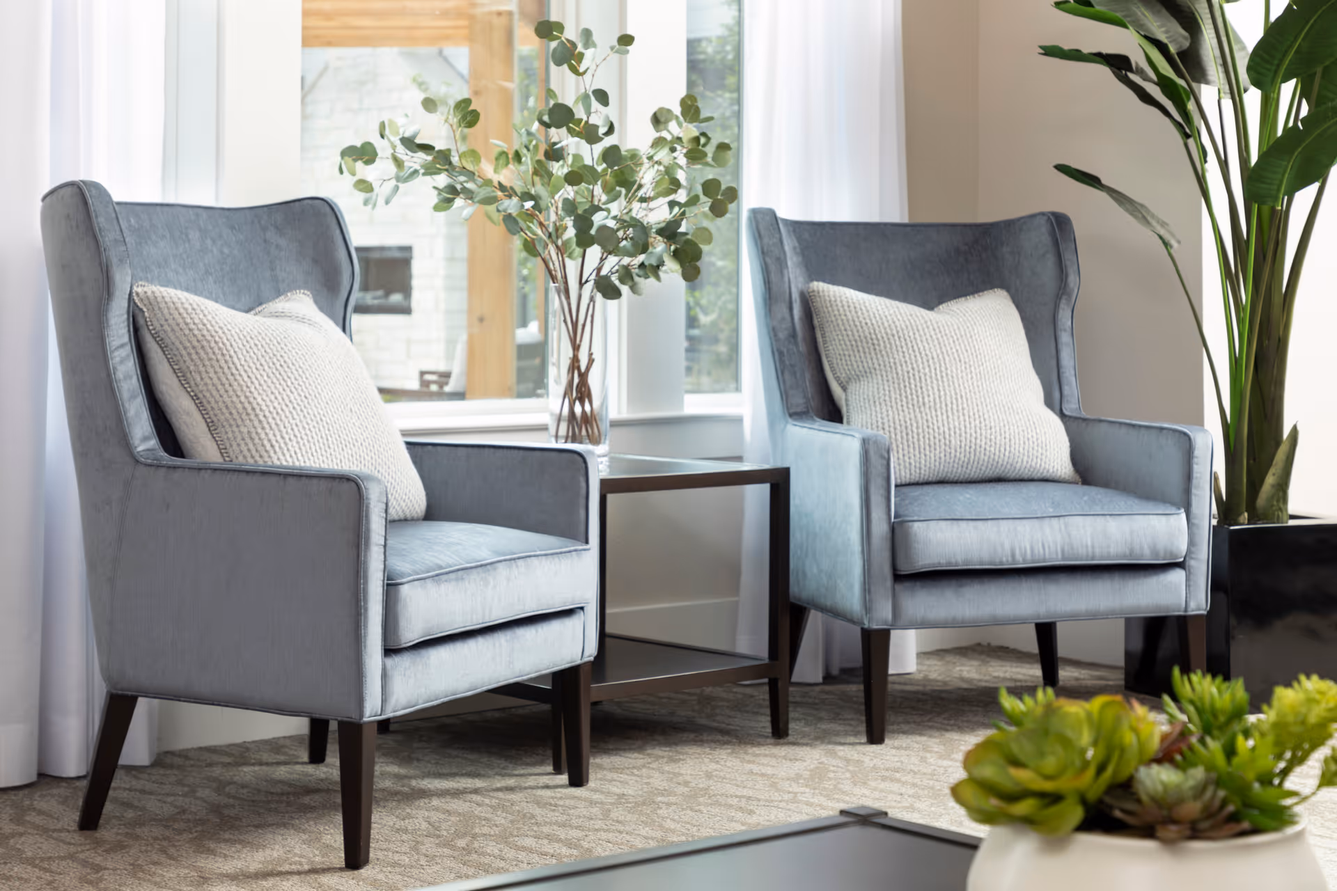 Two gray upholstered armchairs with white knitted pillows are positioned on either side of a small glass-top side table with a vase of green leafy branches. The seating area is set in front of large windows with white curtains, and there is a large potted plant to the right. In the foreground, a table with a white pot containing green succulents is partially visible.
