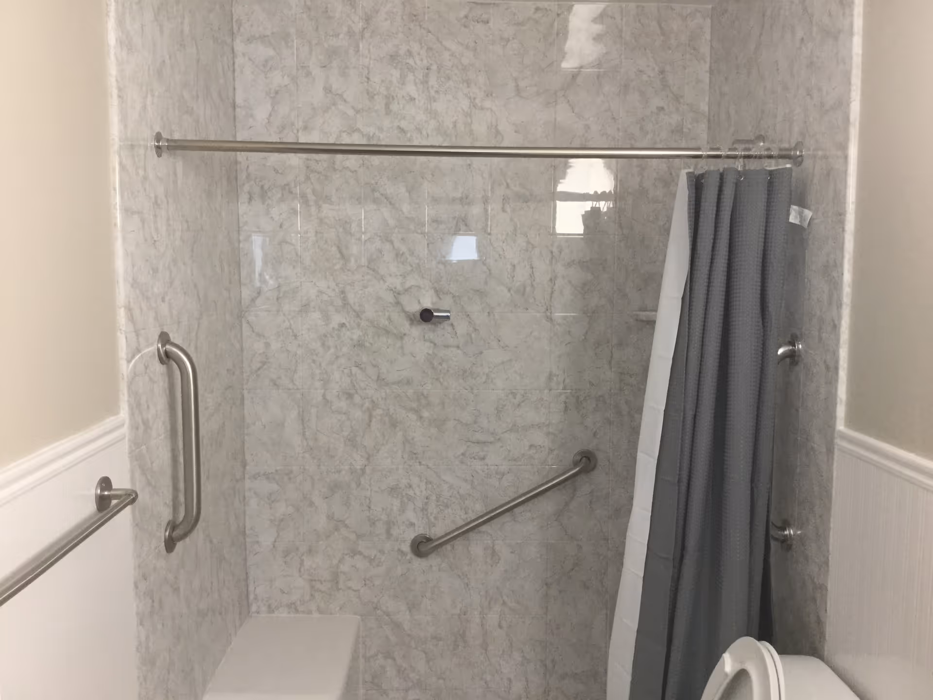 A bathroom shower area with marble-patterned walls, a metal grab bar on the left wall, a diagonal grab bar on the back wall, a shower curtain rod with a gray shower curtain partially drawn, and a white toilet partially visible in the lower right corner.