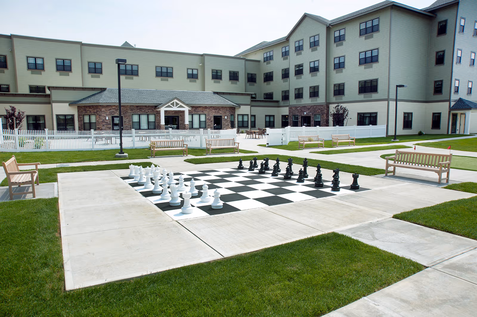 Outdoor courtyard of a senior living facility featuring a large outdoor chessboard, benches, walkways, and the multi-story building in the background.