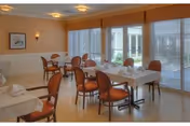 Bright dining room with tables set for meals, orange-upholstered chairs, and large windows with blinds.