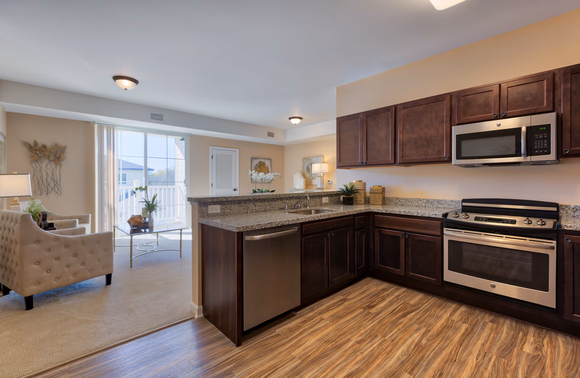 A modern kitchen with dark wood cabinets, granite countertops, stainless steel appliances including a microwave, oven, and dishwasher. The kitchen opens into a living area with beige carpet, a tufted armchair, a glass coffee table with a plant, and a sliding glass door leading to a balcony.