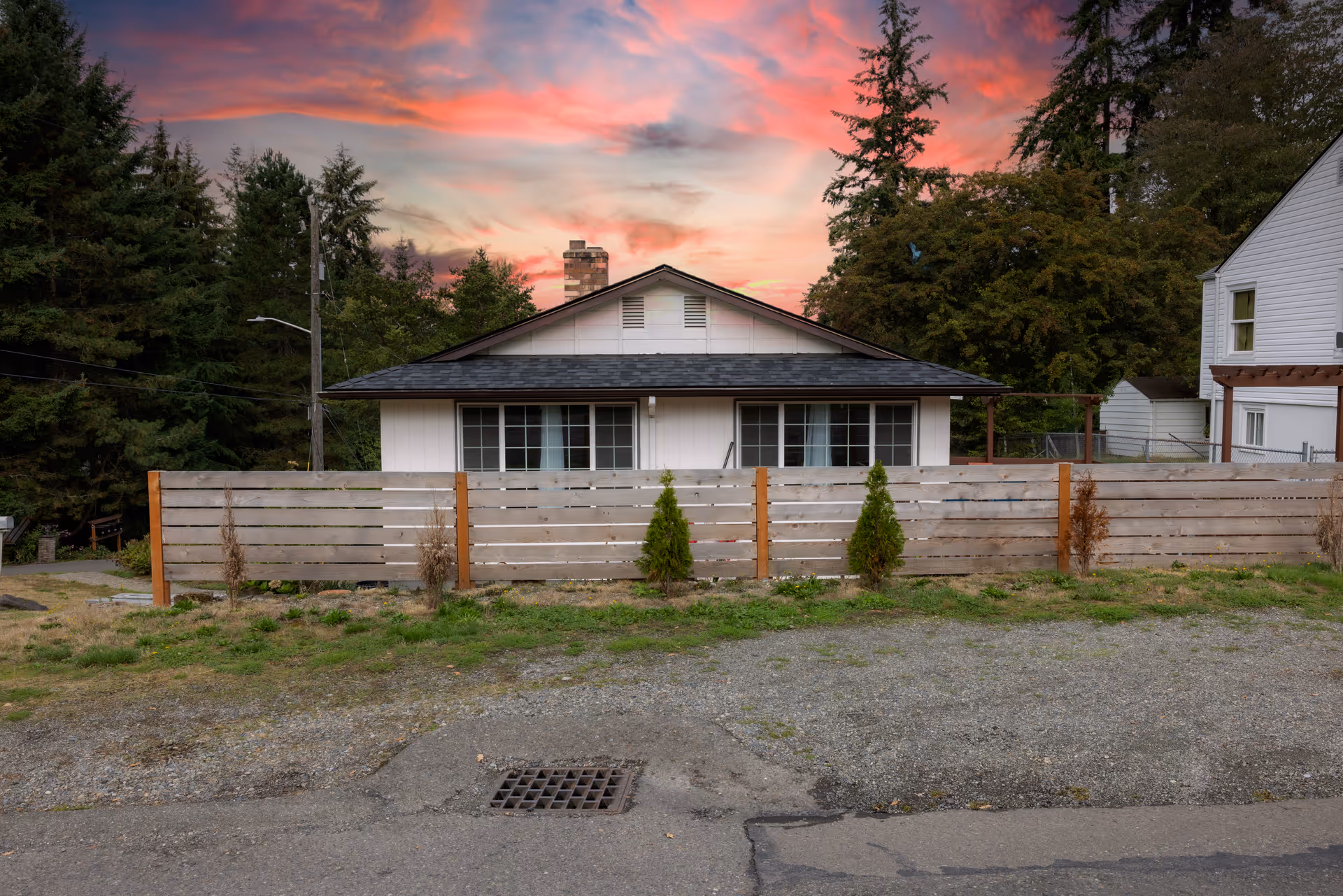 Exterior view of a single-story house with a dark roof and white siding, partially obscured by a wooden fence with small trees planted in front. The house is surrounded by tall evergreen trees under a colorful sunset sky with pink and orange clouds.