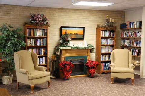 A cozy indoor lounge with two beige armchairs, a fireplace, bookshelves, and poinsettias.