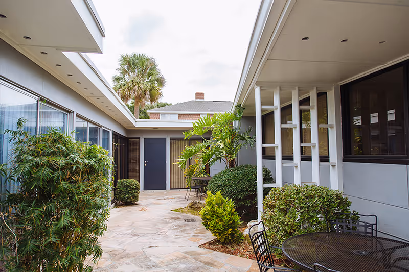 Outdoor courtyard area of Her Heart Village Assisted Living Facility with stone flooring, green plants and bushes, metal tables and chairs, and a palm tree visible in the background under a cloudy sky.