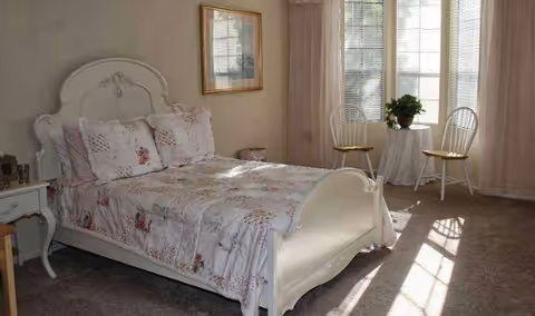 Sunlit bedroom with a white ornate bed dressed in floral bedding and two chairs with a small table by a window.