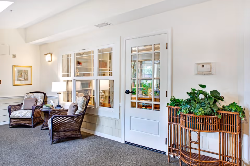 Bright sitting area with two wicker armchairs, a side table and lamp beside a glass-paneled door and a plant stand.