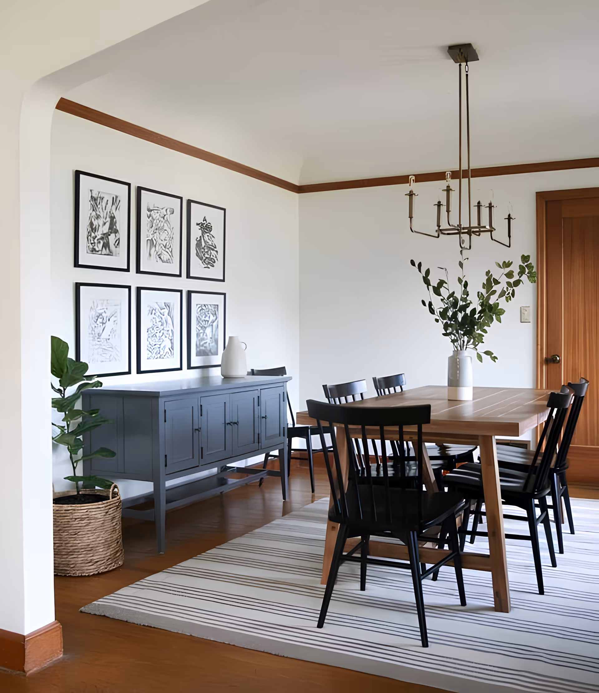 A dining room with a wooden table surrounded by six black chairs on a striped rug. A vase with green leafy branches is placed on the table. Against the wall, there is a blue sideboard with a white pitcher on top and six framed black and white artworks hanging above it. A potted plant in a woven basket is positioned next to the sideboard. The room features wooden trim and a modern chandelier hanging from the ceiling.