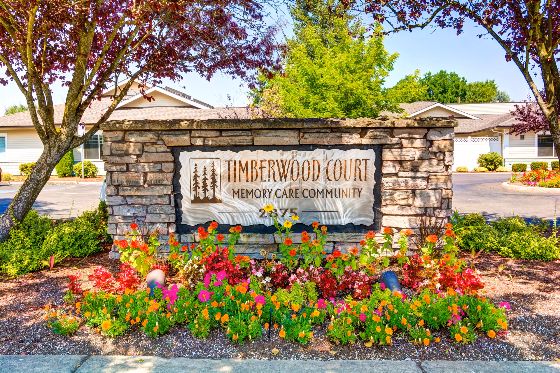 Stone sign for Timberwood Court Memory Care Community surrounded by colorful flowers and trees, with a building and driveway visible in the background.