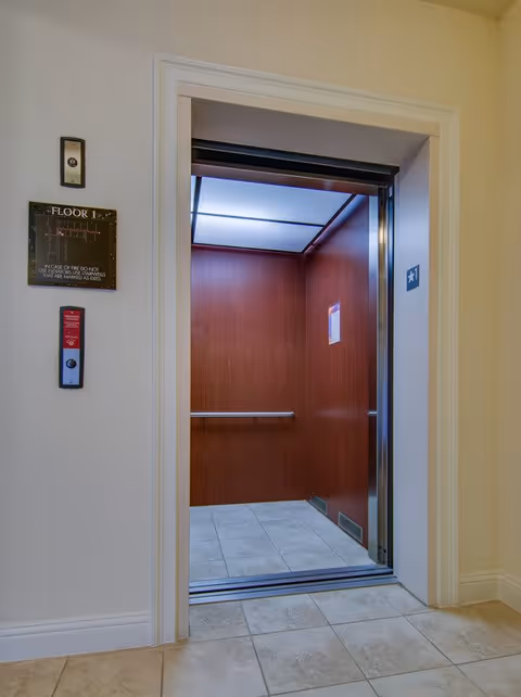 Open elevator with wood-paneled interior walls, tiled floor, and metal handrails inside a building hallway with beige walls and tiled floor.