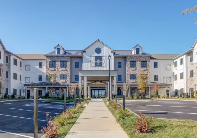 Front exterior view of Mercer Hill at Doylestown, a three-story senior living facility with a combination of stone and white siding, a covered entrance, and a clear blue sky above.