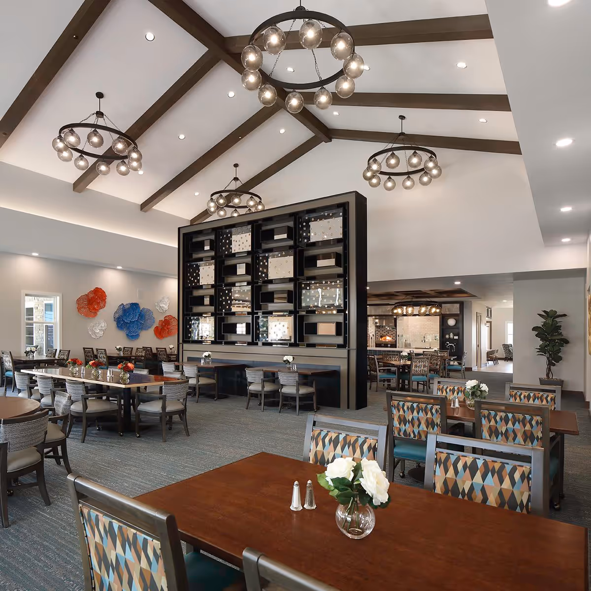 Spacious senior living dining room with multiple wooden tables and chairs featuring colorful patterned upholstery. The room has high vaulted ceilings with exposed wooden beams and modern circular chandeliers. A decorative black shelving unit divides the space, and there are colorful wall decorations on one side. The area is well-lit with recessed lighting and natural light from windows.
