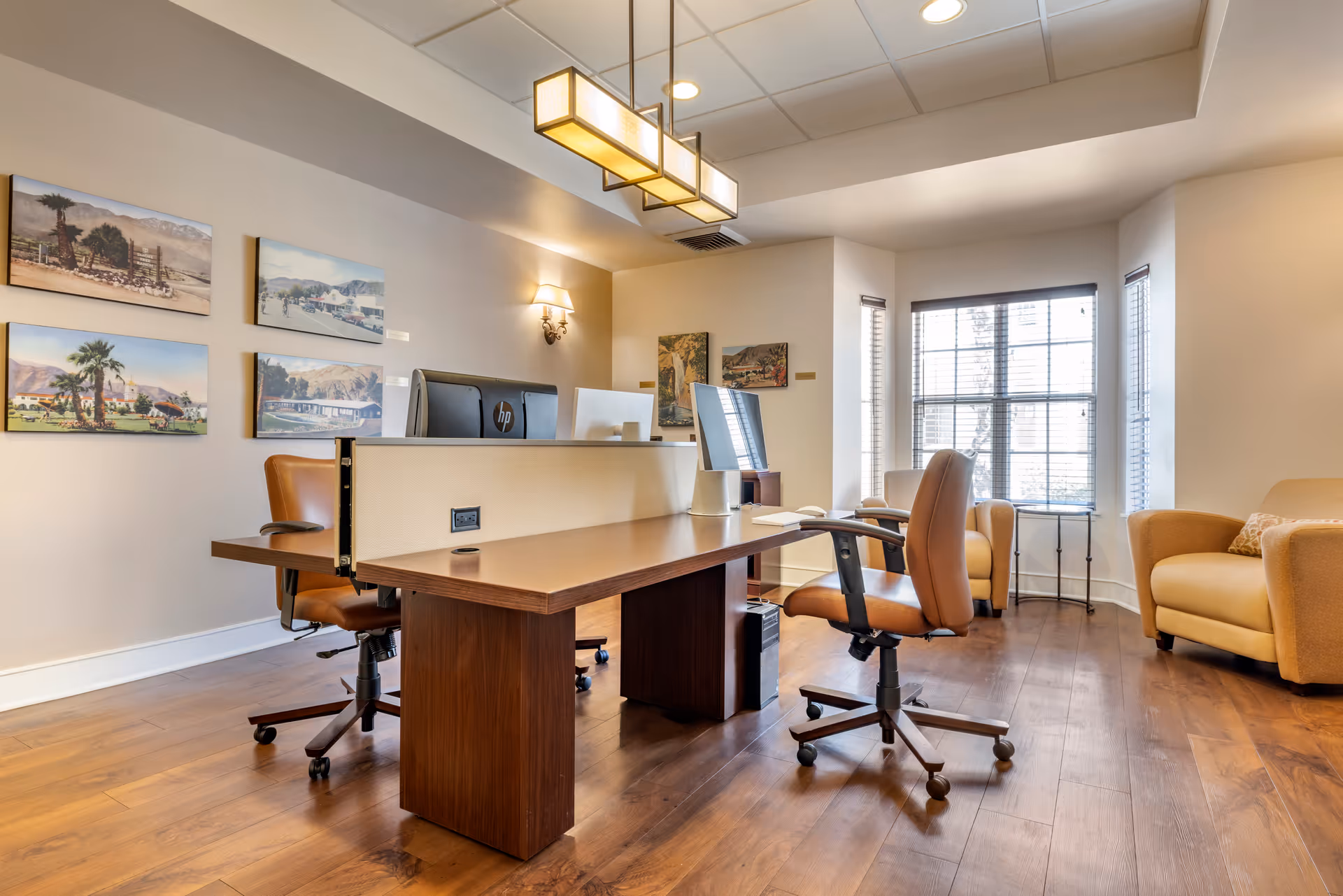 A bright office space with a wooden desk featuring two computer monitors and two brown office chairs on wheels. The room has wooden flooring, beige walls adorned with framed landscape pictures, and a large window with blinds letting in natural light. There are also two beige armchairs near the window and a modern rectangular ceiling light fixture.