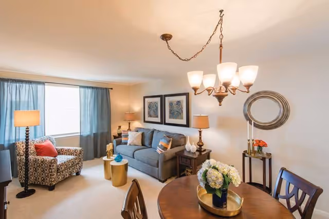 A cozy living room with a gray sofa adorned with decorative pillows, a patterned armchair with a red cushion, two gold side tables, and a floor lamp near a window with blue curtains. The room also features a wooden dining table with chairs, a floral centerpiece, two framed artworks on the wall, a round mirror, and a chandelier hanging from the ceiling.