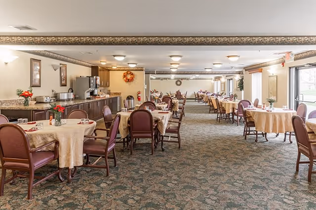 A spacious dining room with multiple round tables covered with beige tablecloths, each surrounded by several chairs with maroon cushions. The room has a patterned carpet and a long counter along one wall with a coffee machine and other appliances. There are framed pictures on the walls and large windows letting in natural light.