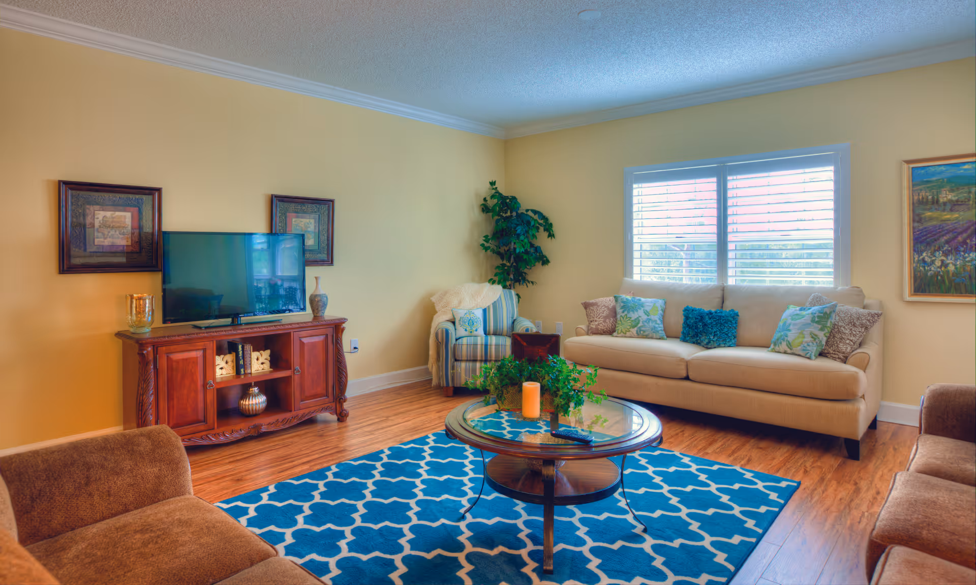 A cozy living room with a beige sofa adorned with colorful pillows, a striped armchair with a throw blanket, a wooden coffee table with a plant and candle, a blue patterned rug, and a wooden TV stand with a flat-screen TV and decorative items. The walls are painted yellow and decorated with framed artwork, and there is a window with white blinds letting in natural light.