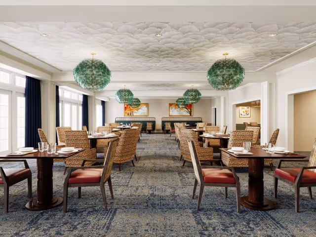 Spacious dining room with multiple round wooden tables, patterned upholstered chairs, large windows, and green hanging pendant lights.