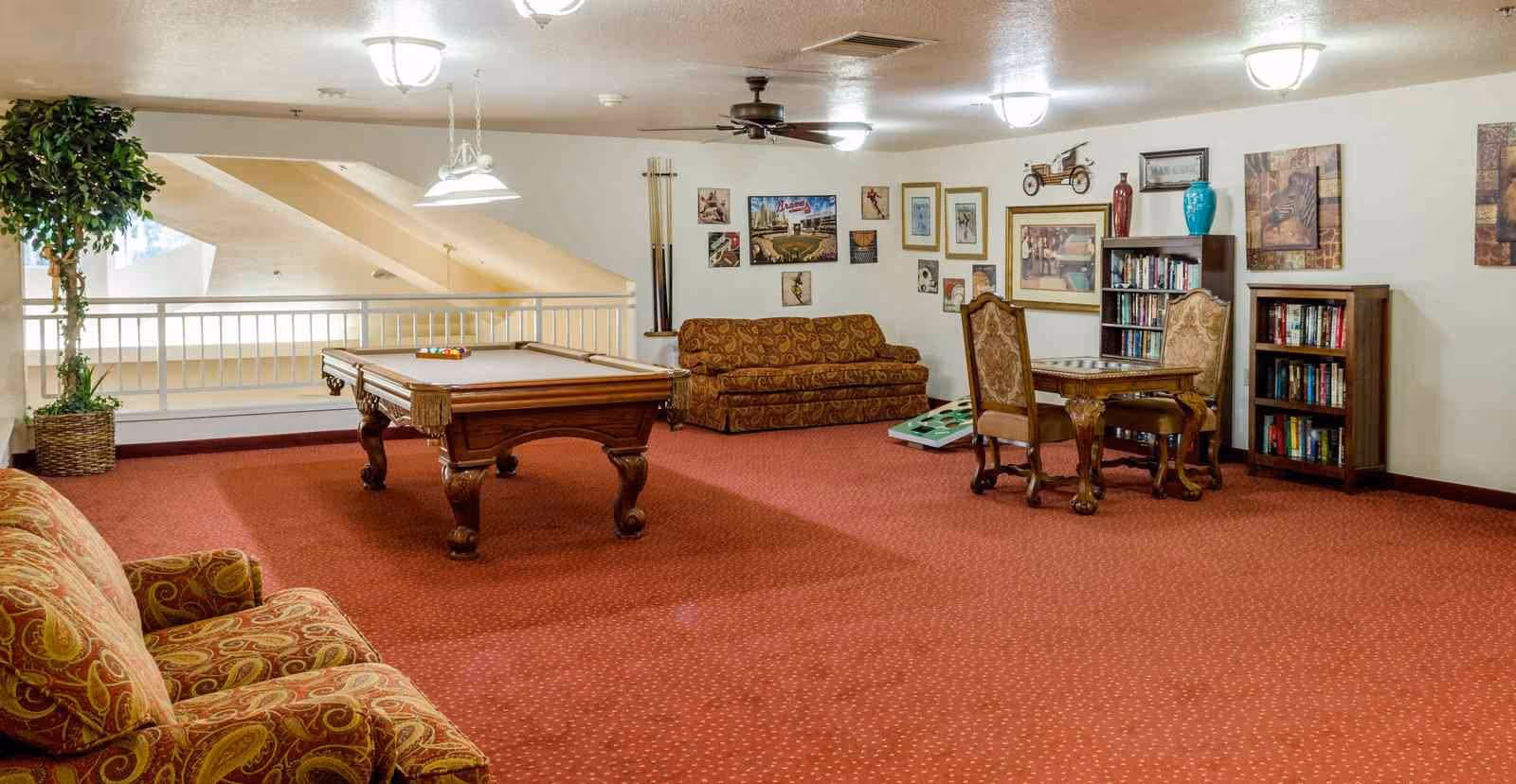 A spacious common room with red carpet featuring a pool table in the center, a patterned sofa and armchair on the left, a small table with two chairs on the right, bookshelves filled with books, framed pictures on the walls, and a potted plant near a railing overlooking a lower level.