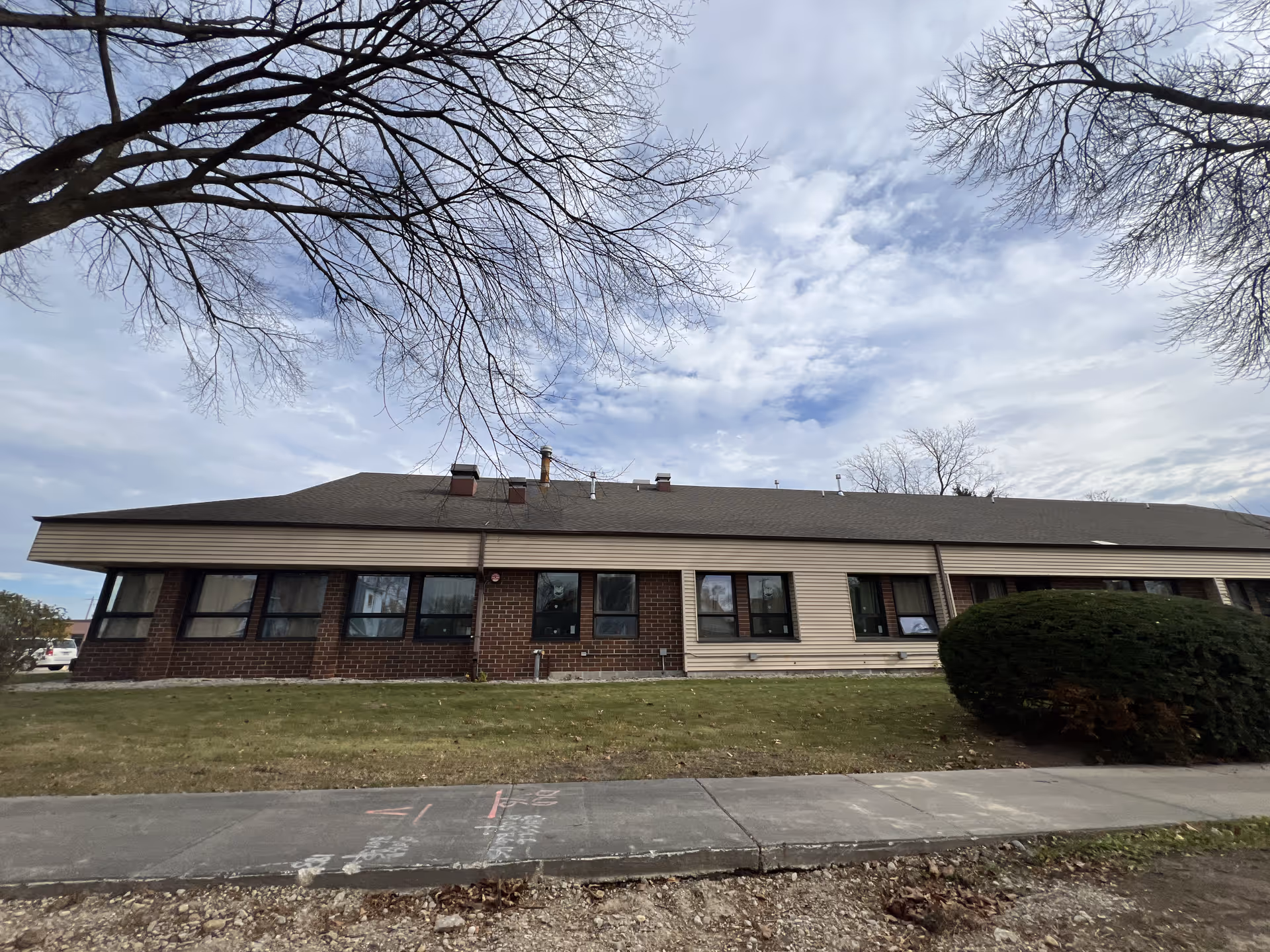 Single-story building with a sloped roof, multiple windows, and a mix of brick and siding exterior. The building is surrounded by a grassy area with a sidewalk in front and leafless trees overhead under a partly cloudy sky.