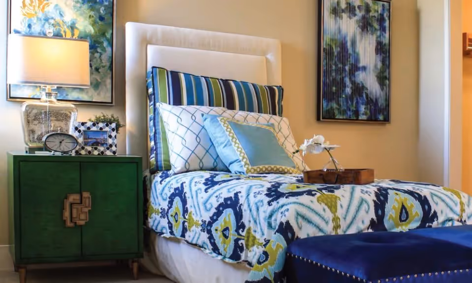 A neatly made single bed with a white upholstered headboard, decorated with multiple pillows in blue, green, and white patterns. A green nightstand beside the bed holds a clear glass lamp, a small clock, a framed photo, and a small plant. Two abstract paintings hang on the beige walls above and beside the bed. A blue upholstered bench is at the foot of the bed.