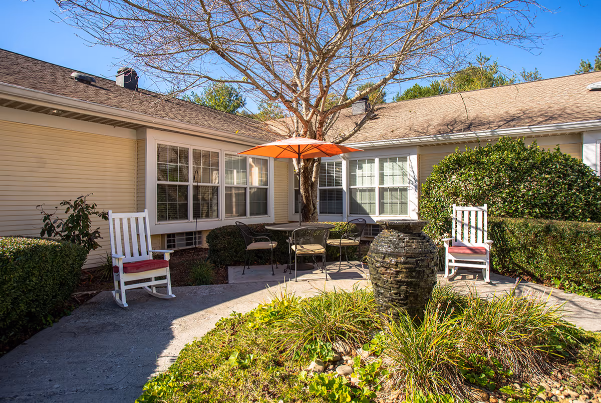 Outdoor patio area at TerraBella Morristown with two white rocking chairs with red cushions, a round table with four chairs under an orange umbrella, surrounded by bushes and a large tree. A stone water fountain is in the foreground with green plants around it.