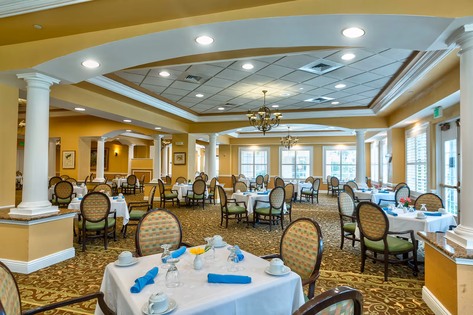 A spacious dining room with multiple round tables covered with white tablecloths, each set with blue napkins, cups, and glassware. The room features patterned carpet, large windows with white blinds, decorative columns, and chandeliers hanging from a coffered ceiling.