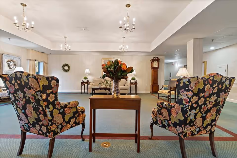 A spacious living room area with two floral patterned armchairs facing a wooden table with a vase of flowers. In the background, there is a beige sofa with side tables and lamps, a grandfather clock, and wall decorations including a wreath and framed art. The room is softly lit with ceiling chandeliers and table lamps.