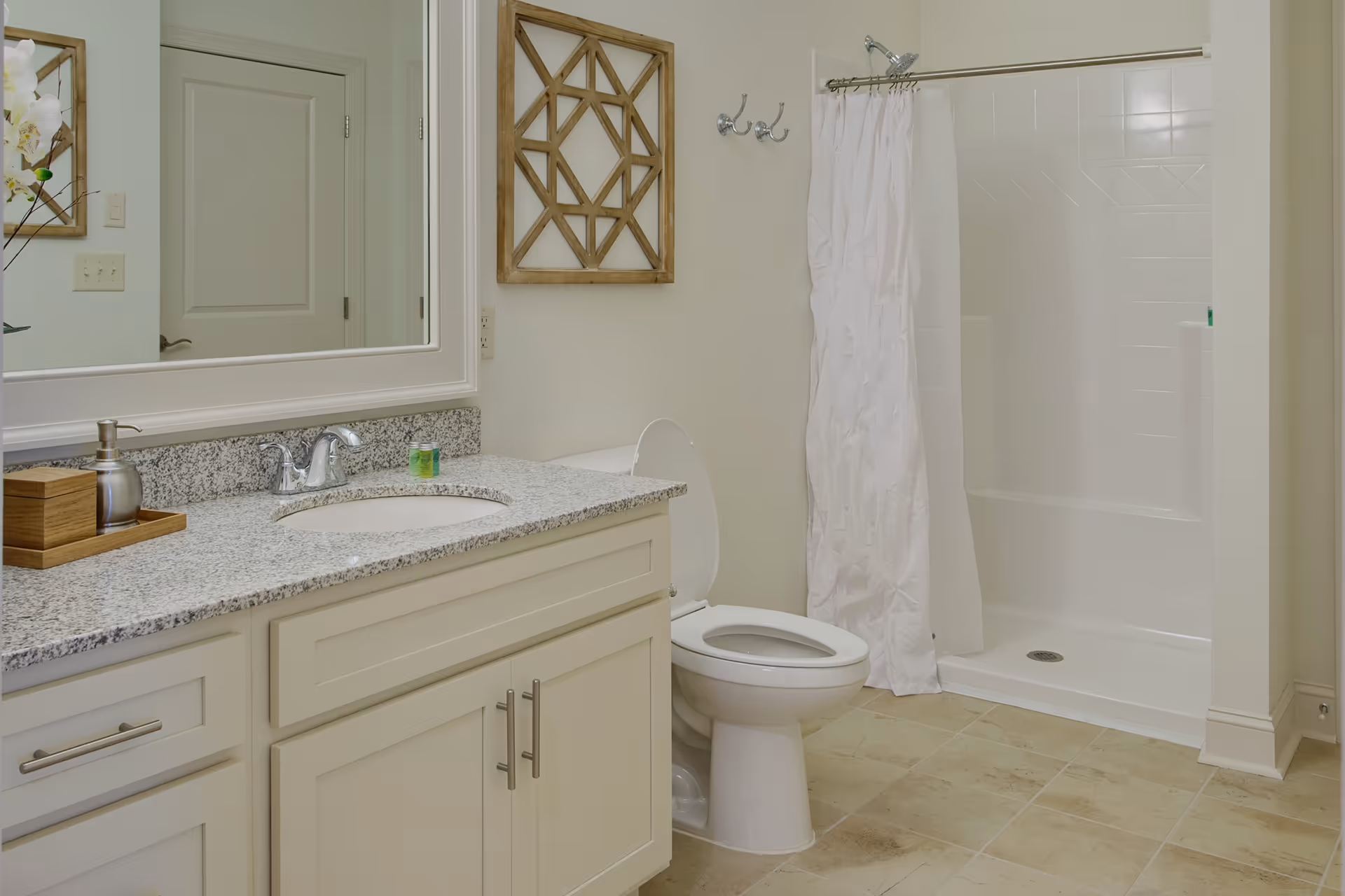 A clean bathroom featuring a white toilet with the lid open, a shower with a white curtain, a granite countertop with a sink, a soap dispenser, and a wooden box. There is a large mirror above the sink and a decorative wooden wall hanging next to the shower.