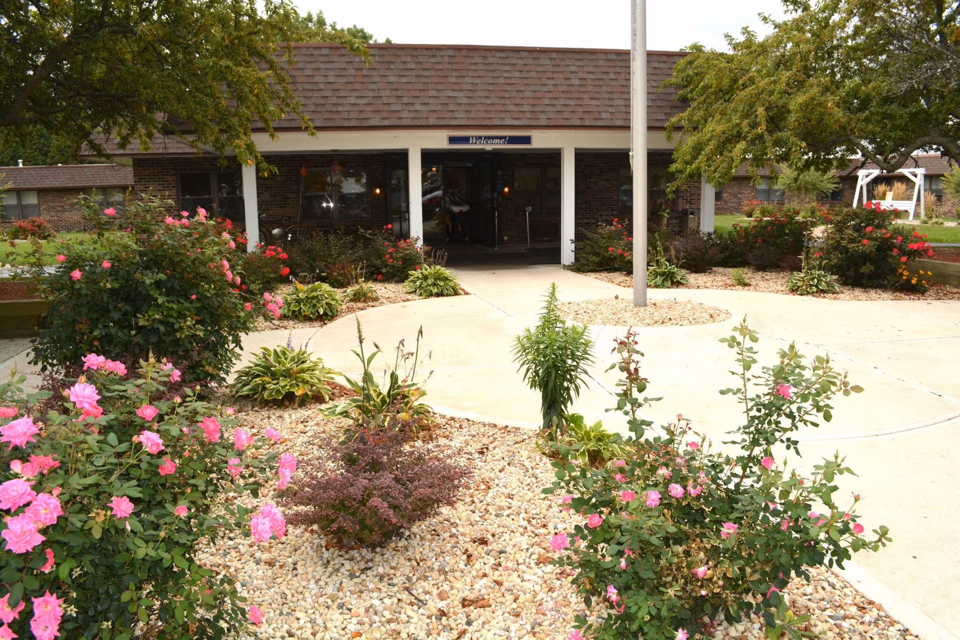 Front entrance of a care facility with a circular driveway, flagpole, and landscaped flower beds with pink roses.