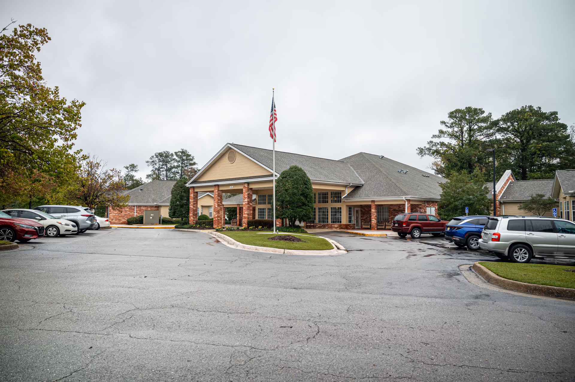 Exterior view of The Landings of Northport senior living facility showing a single-story building with a covered entrance, an American flag on a flagpole in front, surrounded by parked cars and trees under a cloudy sky.