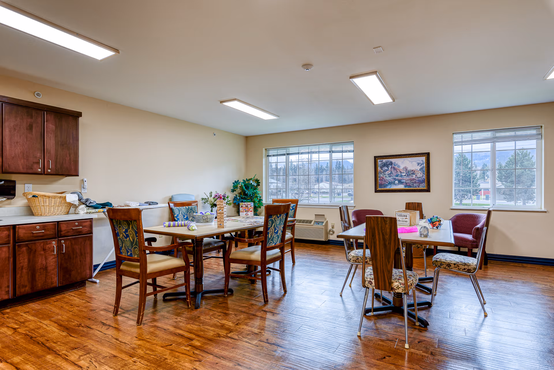 A bright room with wooden flooring and two tables surrounded by chairs. The tables have various items on them including a Jenga game, flowers, and a welcome sign. There are two large windows letting in natural light, a framed painting on the wall, and a cabinet with a basket and folded towels on the left side.