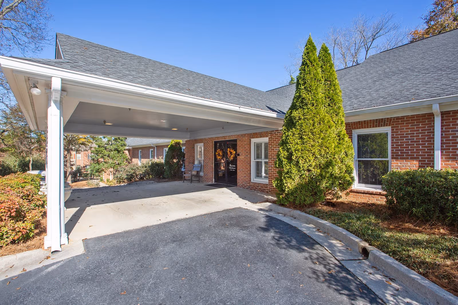 Entrance area of Bethel Gardens Assisted & Memory Care facility showing a covered drop-off zone with brick exterior walls, windows, a door decorated with wreaths, and surrounding greenery including bushes and a tall conical tree under a clear blue sky.