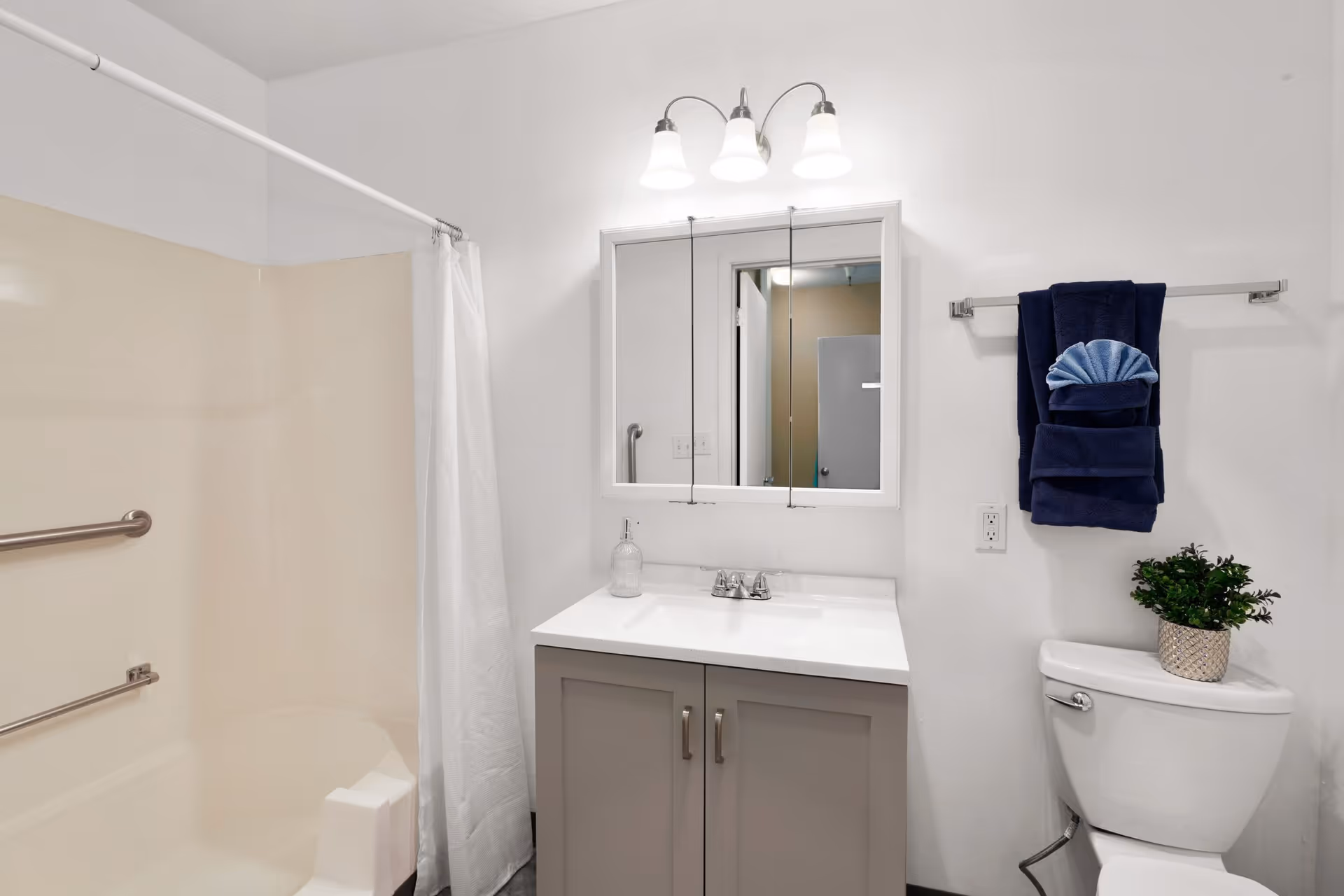 A clean and modern bathroom featuring a beige bathtub with a white shower curtain, a vanity with a white countertop and gray cabinet, a three-light fixture above a mirrored medicine cabinet, a toilet with a small potted plant on top, and a towel rack holding neatly folded dark blue and light blue towels.