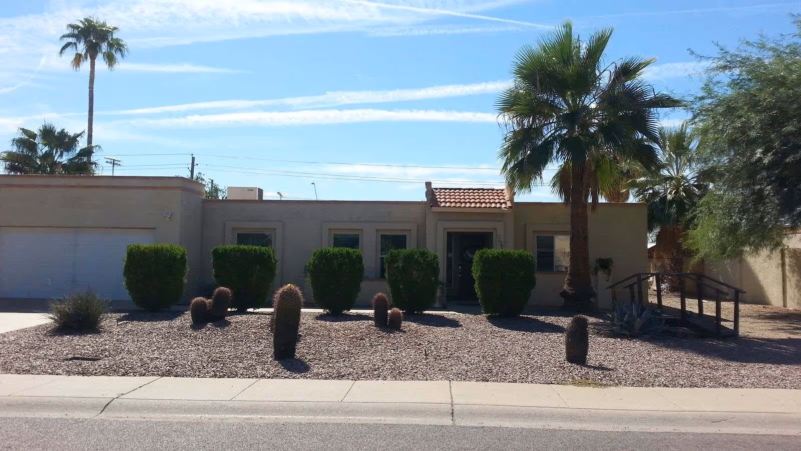 Single-story stucco building front with a gravel desert yard, cacti, trimmed shrubs, and palm trees under a clear blue sky.