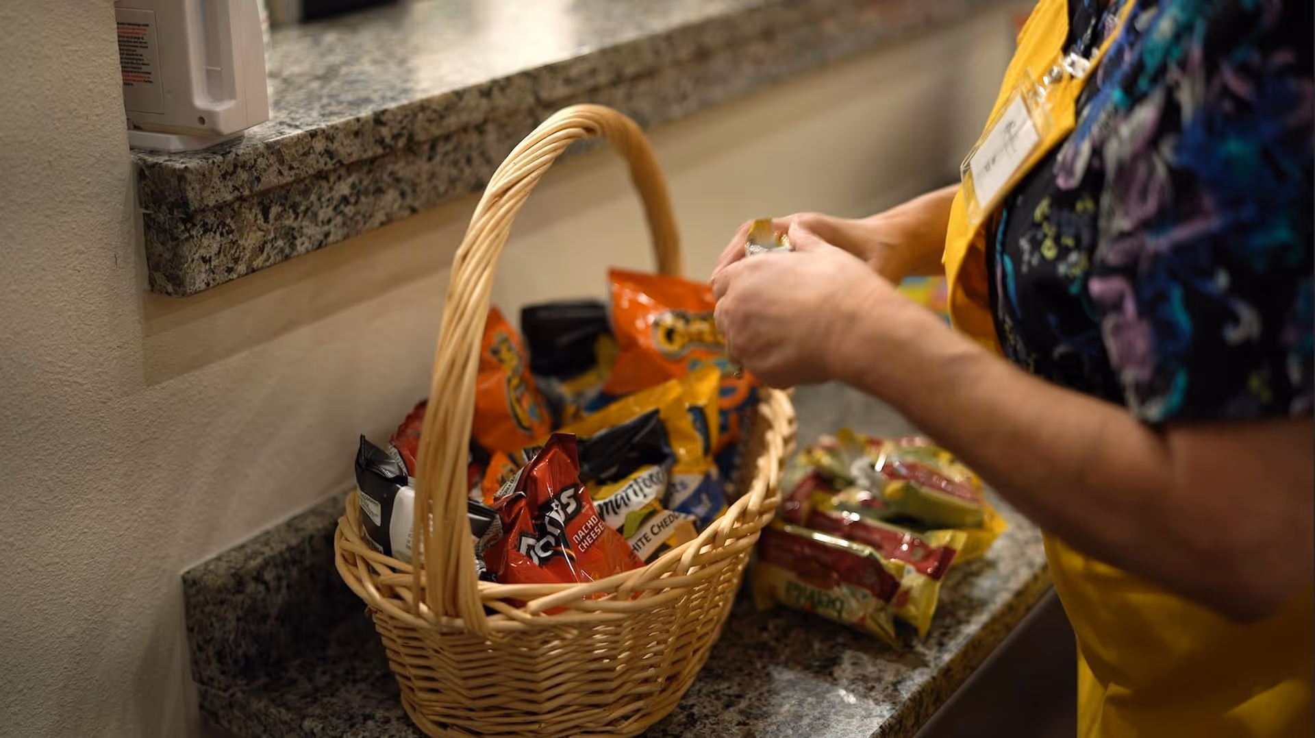 A person wearing a yellow apron is placing snack bags into a wicker basket on a granite countertop. Various snack bags are visible both inside the basket and on the counter.