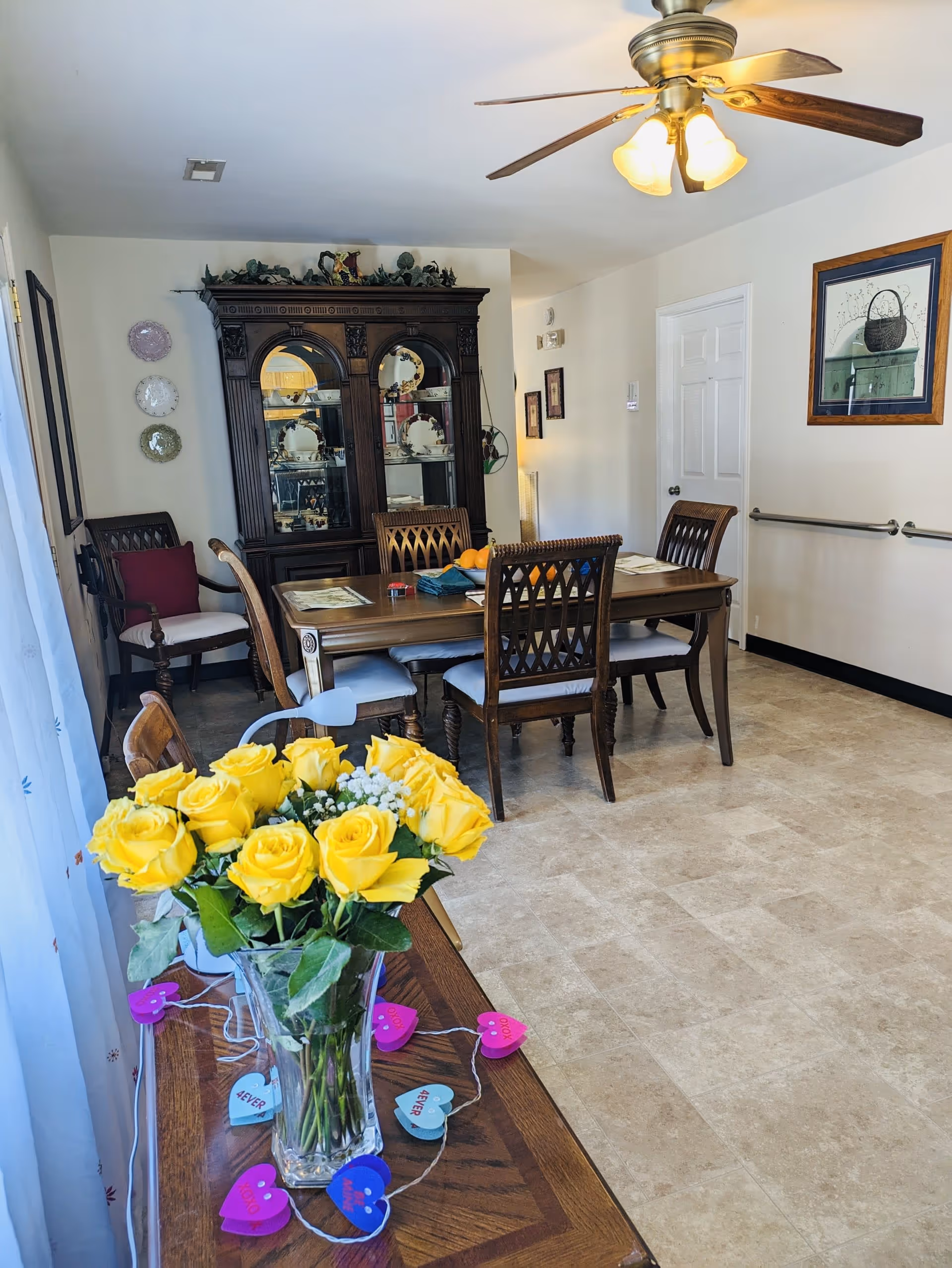 A dining room with a wooden dining table and six chairs. A dark wooden china cabinet with decorative plates is against the far wall. A ceiling fan with lights is mounted on the ceiling. A vase with yellow roses is placed on a wooden side table in the foreground, decorated with a string of colorful heart-shaped lights. The room has light-colored tiled flooring and light walls with framed artwork and decorative plates.