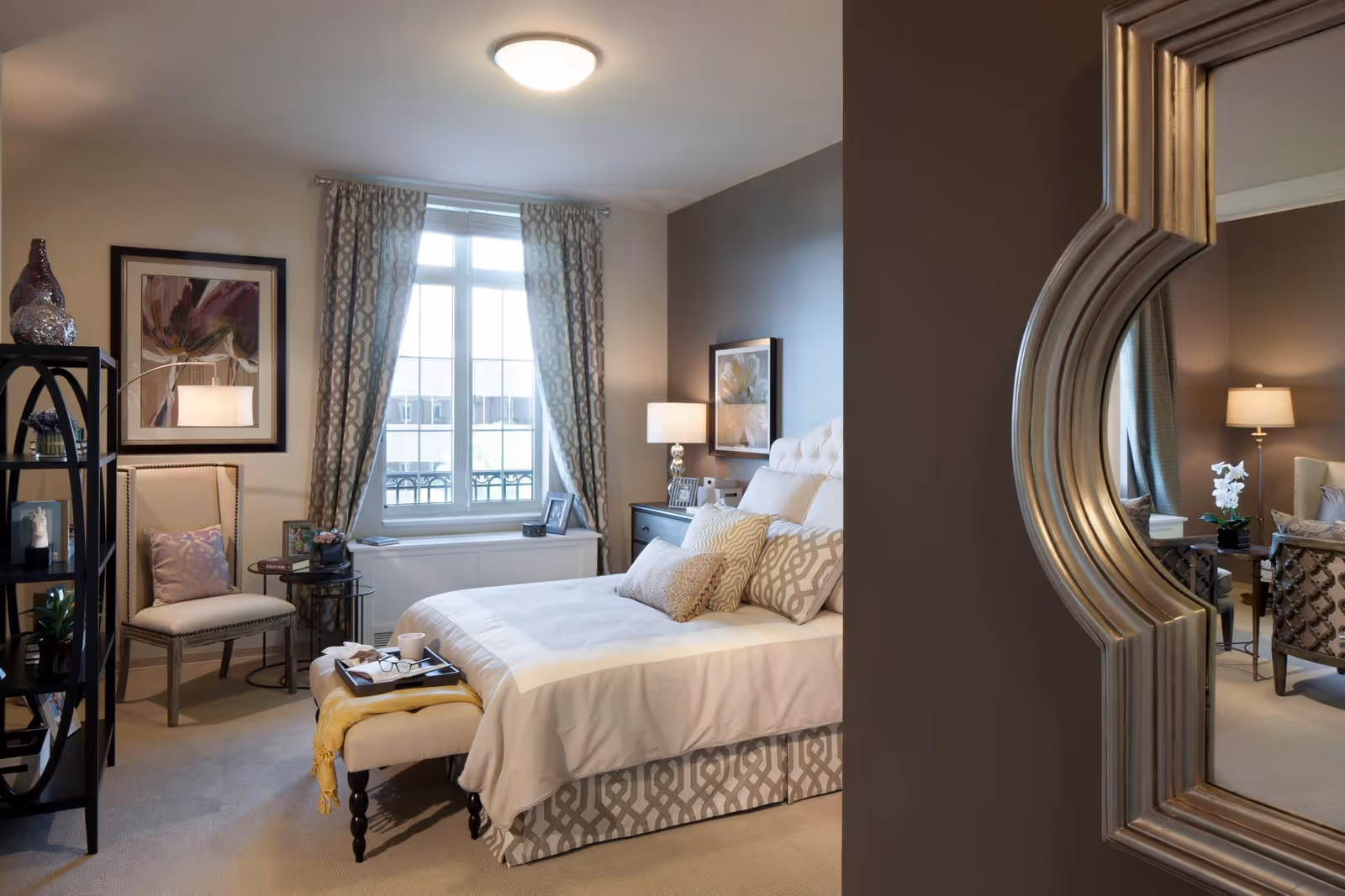 A cozy and well-decorated bedroom in a senior living facility featuring a bed with multiple pillows and a patterned bed skirt, a cushioned bench at the foot of the bed with a tray holding a cup and glasses, a chair with a pillow, a side table, a window with patterned curtains, framed artwork on the walls, and a large decorative mirror reflecting part of a sitting area with chairs and a lamp.